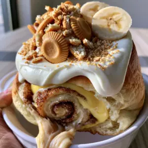 A handheld smartphone photo showing a gooey banana pudding cinnamon roll being lifted, revealing a thick dough stretch and vanilla pudding filling. (banana pudding cinnamon rolls)